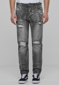 Jeans in denim grigio sbiadito con strappi usurati e bordi sfrangiati, caratterizzati da una vestibilità standard e un design a cinque tasche, indossati con scarpe da ginnastica nere.