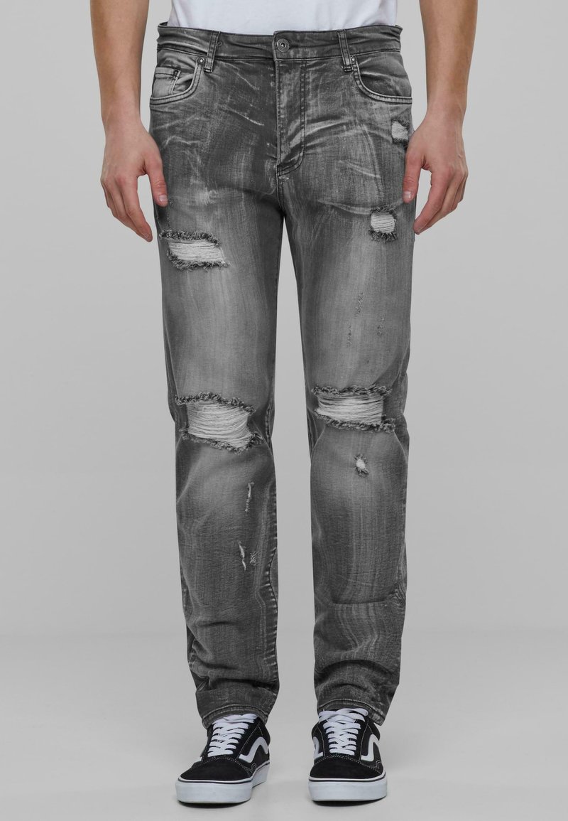 Jeans in denim grigio sbiadito con strappi usurati e bordi sfrangiati, caratterizzati da una vestibilità standard e un design a cinque tasche, indossati con scarpe da ginnastica nere.