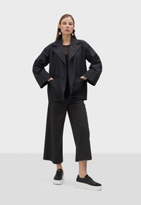 Černá, lehká bunda s širokými klopami a předními kapsami, v kombinaci s černým texturovaným topem, širokými kalhotami a platformovými černými teniskami.