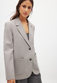 Grey blazer with a tailored fit, featuring a notched lapel, two front pockets, and two black buttons. Smooth texture and structured design.