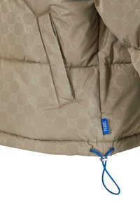 Beige pufferjakke med et geometrisk mønster, der har en blå detaljeringsetiket og snor i kanten. Glat tekstur med quiltede sektioner.