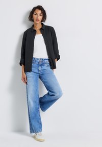 Donna appoggiata al muro che indossa una camicia nera con bottoni, una maglietta bianca, jeans a gamba larga blu chiaro e sneakers bianche.