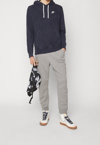 Marinblå huvtröja med vita snören och logotyp, gråa träningsbyxor och vita höga sneakers med bruna sulor. Svart och gråmönstrad väska.