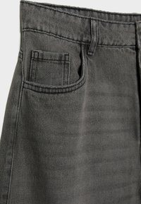 Acercamiento a un bolsillo de pantalón de mezclilla gris con costuras detalladas y un ojal para cinturón sobre un fondo liso.