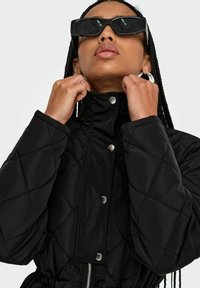 Veste matelassée noire avec un col montant, des boutons pression et une taille cintrée. Le mannequin porte des lunettes de soleil noires surdimensionnées et des boucles d'oreilles en argent.