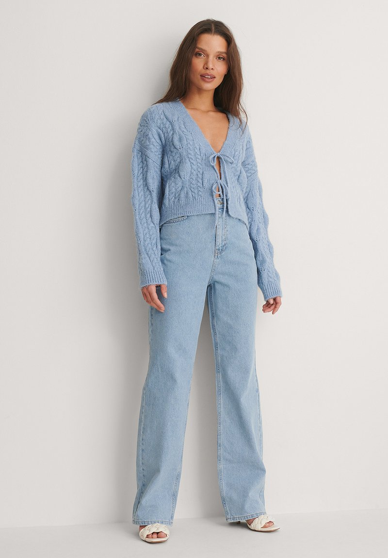 Gilet court en tricot torsadé bleu avec fermeture à nouer, associé à un jean flare en denim clair taille haute et des sandales blanches à lanières.