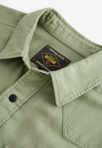 Green cotton shirt featuring a pointed collar, black snap button, and a tag with 'Premium Grade' and brand logo. Textured fabric detail visible.