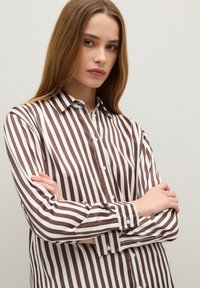 Young woman with long blonde hair wearing a brown and white vertical striped button-up shirt, arms crossed in front of a plain light background.
