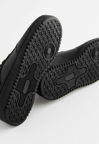 Semelles de baskets noires avec une surface en caoutchouc texturée, présentant un motif circulaire et hexagonal ainsi qu'un accent de design central.