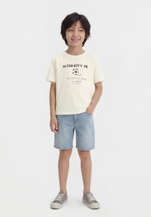 Chłopiec stojący z jedną ręką w kieszeni dżinsowych szortów, ubrany w kremowy t-shirt z piłką nożną i napisem oraz szare tenisówki, lekko się uśmiecha.