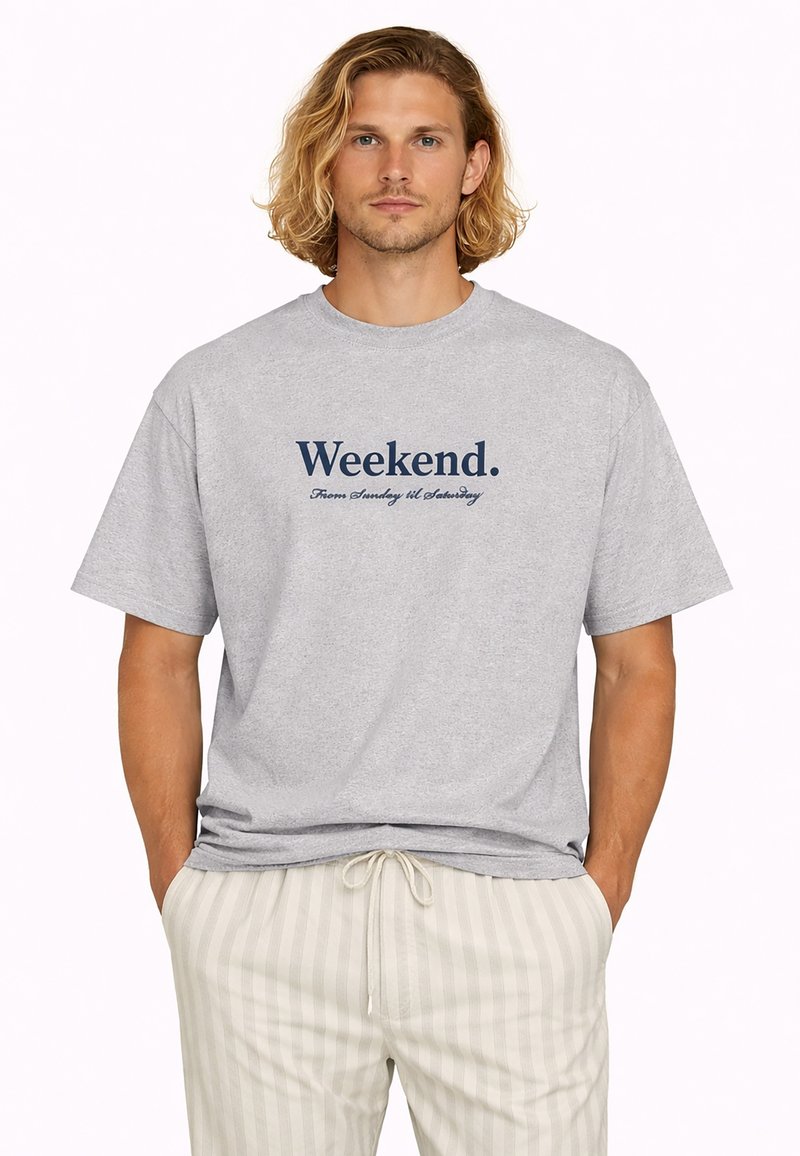Homme blond aux cheveux mi-longs portant un T-shirt gris avec le texte « Week-end. Du dimanche au samedi » et un pantalon léger à rayures, les mains dans les poches.