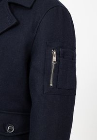 Granatowy wełniany płaszcz z usztywnionym kołnierzem, z kieszenią na suwak na rękawie oraz dużym zapięciem na guziki z przodu. Teksturowana tkanina.