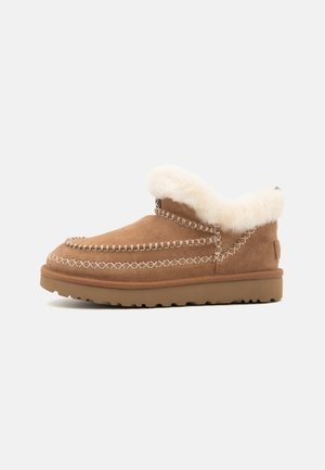 Zapato de ante marrón tipo slip-on con forro de pelo sintético blanco, costuras en contraste y suela de goma. Diseño sencillo y casual adecuado para uso en interiores.