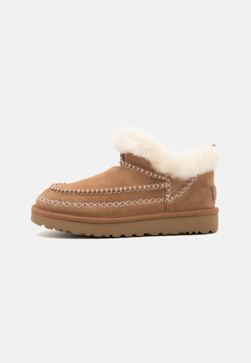 Zapato de ante marrón tipo slip-on con forro de pelo sintético blanco, costuras en contraste y suela de goma. Diseño sencillo y casual adecuado para uso en interiores.