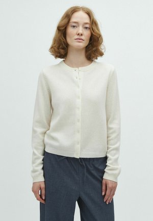 Femme aux cheveux bouclés mi-longs, portant un cardigan crème boutonné et un pantalon large foncé, sur un fond clair uni.