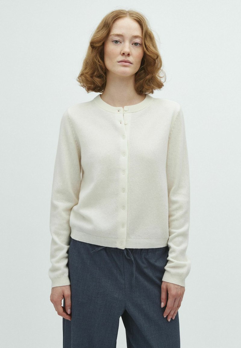 Femme aux cheveux bouclés mi-longs, portant un cardigan crème boutonné et un pantalon large foncé, sur un fond clair uni.