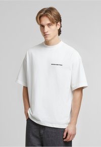Weißes Baumwoll-T-Shirt mit kurzen Ärmeln, Rundhalsausschnitt und schwarzem "VERBOTEN"-Text im linken Brustbereich. Klassische Passform, glatte Textur.