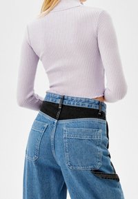 Mujer que lleva un cuello de tortuga acanalado de color púrpura claro y jeans de tiro alto azul con parches negros y azul oscuro en la cinturilla y los bolsillos.