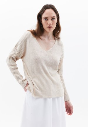 Beige gestrickter Pullover mit V-Ausschnitt, gerippten Ärmeln und seitlichen Schlitzen. Die Textur ist weich und schimmert leicht vor einem weißen Hintergrund.