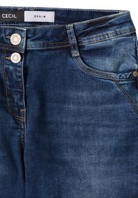 Jeans in denim blu con una texture liscia, tasche frontali e due bottoni metallici in vita. L'etichetta riporta "CECI" e "DENIM."