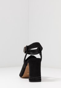 Svart mocka högklackad sko med vristrem och gyllene spänne, tjock klack och vitt stygn på klackens och remmens kanter.