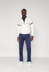 Hvit zip-up jakke med marineblå og oransje striper, kombinert med marineblå bukser med merke-logo. Modellen har på seg hvite joggesko. Nøytral bakgrunn.