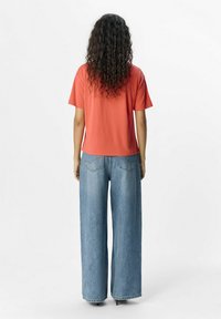 Rød T-shirt med korte ærmer og afslappet pasform, parret med bred-lags blå jeans, der har baglommer og falmet tekstur, vist fra ryggen.