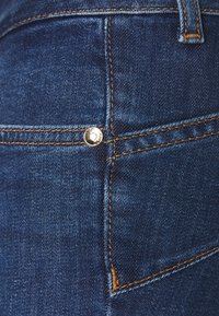 Tessuto di denim blu scuro con cuciture gialle dettagliate e un piccolo rivetto argentato sull'angolo di una tasca posteriore dei jeans.