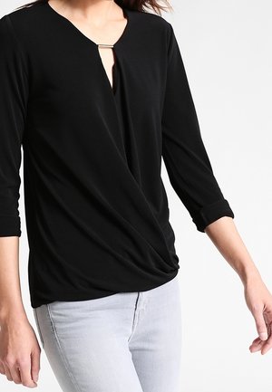 Frau trägt ein schwarzes langärmeliges Oberteil mit gerafftem Vorderteil und silberner Schnalle nahe dem Halsausschnitt, kombiniert mit hellgrauen Hosen.