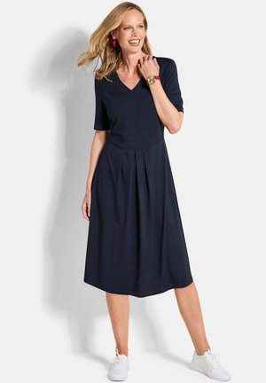 Frau lächelt, trägt ein dunkelblaues V-Ausschnitt-Kleid mit Falten, weiße Turnschuhe, eine rote Uhr und rote Creolen, steht vor einem weißen Hintergrund.