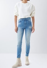 Vaqueros de tiro alto azul claro con un ajuste ajustado, combinados con un sudadera color crema y botines beiges, con un clásico diseño de cinco bolsillos.