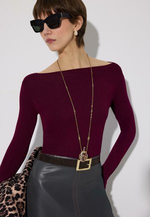 Naine, kes kannab tumedaid päikeseprille, bordoopunast paadi-kaelusega tuba, nahast seelikut laiade vöökohaga, pikka ripatsi kaelakeed ja hoiab käes leopardi mustriga mantlit.