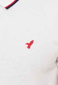 Άσπρο πόλο μπλουζάκι με κόκκινο κεντημένο κολιμπρί στο στήθος και γιακά διακοσμημένο με κόκκινες και μπλε ρίγες.