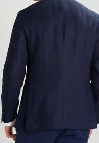 Man klädd i en mörkblå strukturerad kavaj och matchande byxor, sedd bakifrån mot en enkel ljus bakgrund.