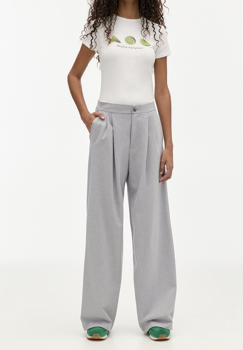 Femme portant un t-shirt blanc avec des illustrations de citron vert et du texte, un pantalon gris à jambes larges et des baskets vertes, mains relâchées le long du corps.