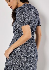 Navy Kleid mit floralen weißen Mustern, kurzen puffärmeln, gerafftem Taillenbereich und Rüschendetails am Ausschnitt. Leichte Stoffqualität mit weicher Textur.