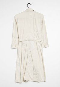 Langes, hellbeiges Kleid aus Baumwolle. Verfügt über einen Kragen, lange Ärmel, eine taillierte Taille und einen plissierten Rock. Mit Knöpfen an den Ärmeln.