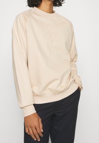 Sudadera beige con cuello redondo, puños y dobladillo acanalados, con un logo "HUGO" en relieve en el frontal. Tejido suave y texturizado.