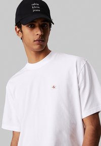 T-shirt in cotone bianco con maniche corte e colletto rotondo, caratterizzato da un piccolo patch del logo rosso e bianco sul lato sinistro del petto. Cappellino nero con testo del marchio.