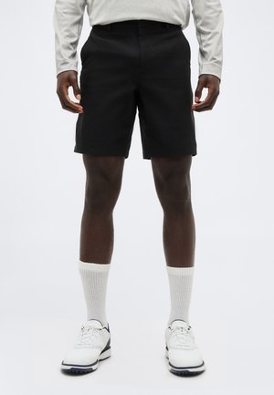 Hombre vestido con pantalones cortos negros, calcetines blancos hasta la mitad de la pantorrilla, zapatos blancos y negros, y una camisa de manga larga gris claro, de pie sobre un fondo blanco.