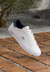 Balti odiniai sportbačiai su tamsiai mėlynais akcentais, rišamosios juostos dizainas ir mažas Lacoste logotipas raudonai ir tamsiai mėlynai. Guminė padas su plokščiu profiliu.