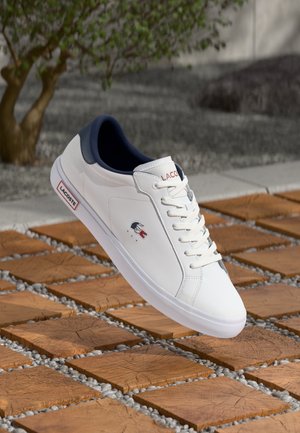Zapatilla blanca de cuero con acentos en azul marino, diseño con cordones y un pequeño logo de Lacoste en rojo y azul marino. Suela de goma con perfil plano.