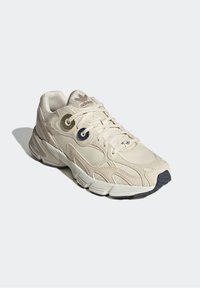 Beige atletiska skor med en överdel av texturerat läder och mesh, runda öglor och en mönstrad gummisula. Utsmyckad med en logotyp på tungan.