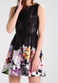 Abito nero senza maniche con corpetto aderente e gonna plissettata; presenta motivi floreali colorati all'orlo in tonalità di rosa, giallo e viola.