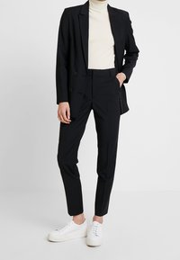 Schwarzer maßgeschneiderter Anzug mit einem doppelt geknöpften Blazer und schmal geschnittenen Hosen, kombiniert mit einem cremefarbenen Rollkragenpullover und weißen Sneakers. Minimalistisches Design.