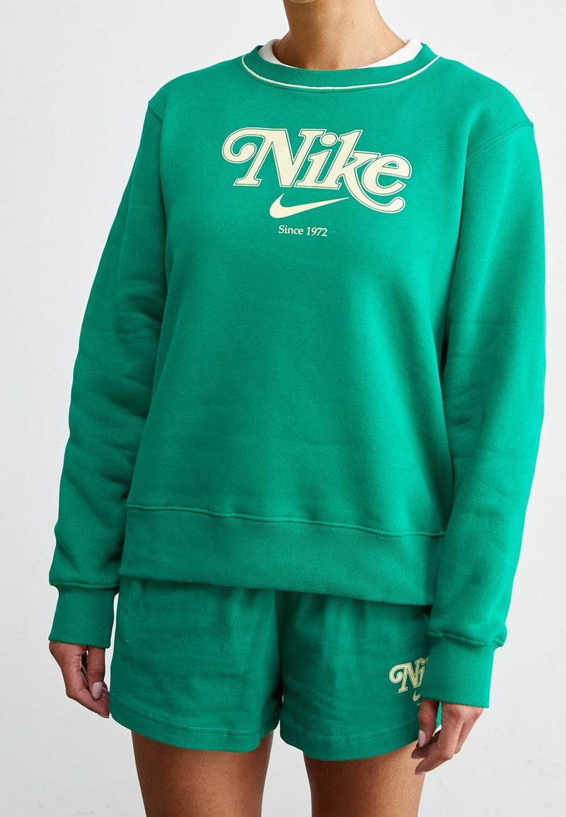 Grön sweatshirt med långa ärmar och ribbad krage; pryds av elfenbensfärgad Nike-text och taglinen "Sedan 1972". Matchas med gröna shorts.