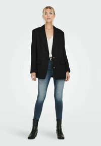 Blazer nero oversize con due tasche anteriori, chiusura a bottoni e rever a forma di punta, indossato sopra una maglietta bianca e jeans skinny blu con dettagli strappati.