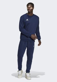 Mørkeblå Adidas sweatshirt og træningsbukser i blødt stof, med ribbede manchetter, rund hals og små logo detaljer. Skoene er hvide med guld accenter.