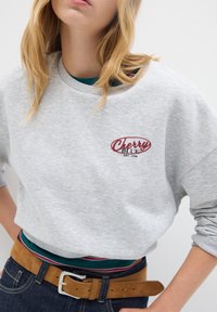 Donna che indossa una felpa grigia chiara con il logo "Cherry Blue EST. 1998", sovrapposta a una maglietta a righe, jeans blu e una cintura in suede marrone.