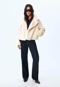 Creme farvet faux fur cropped jakke med store klapper, knaplukning og sidelommer; parret med mørk denim brede bukser og spidse sko.